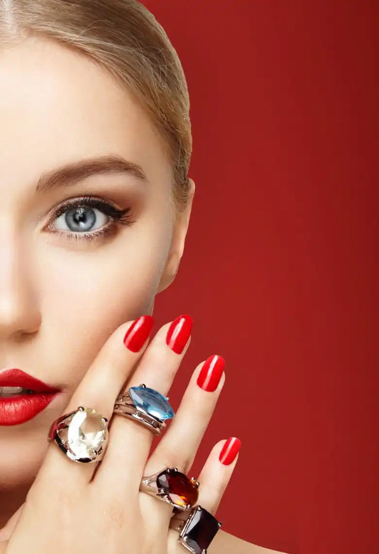 A fair-skinned woman with blonde hair, blue eyes, red lipstick, and red nail polish, wearing several large, colorful gemstone rings.