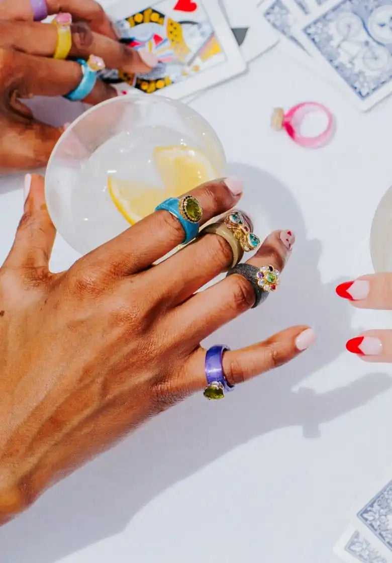 Hands adorned with colorful, gem-studded rings, holding a drink with a lemon slice.